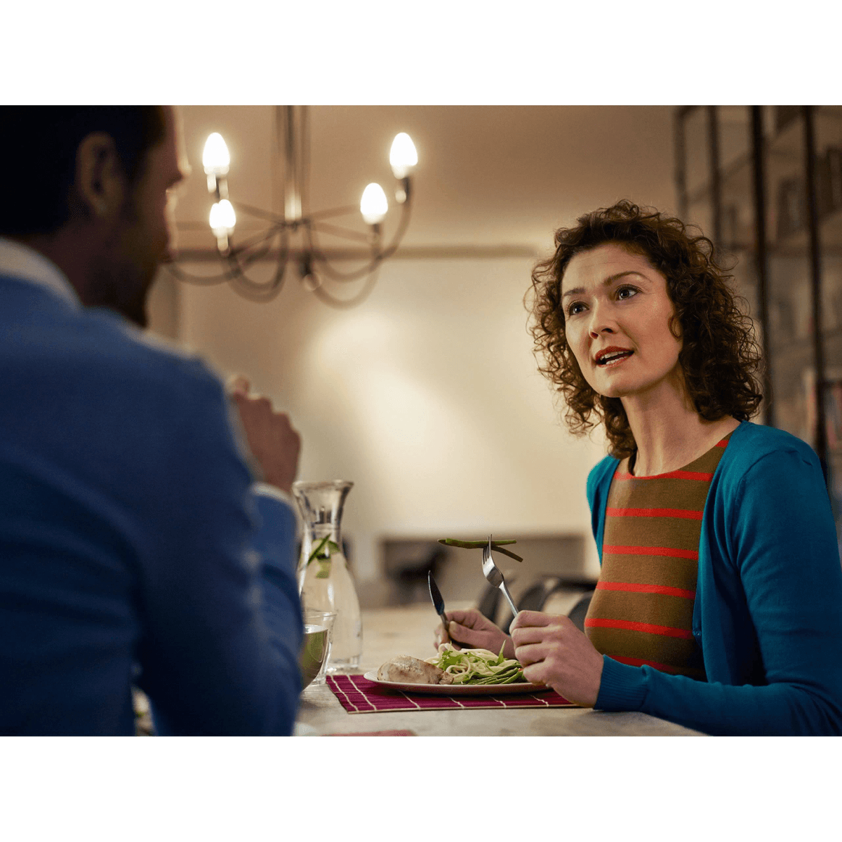 Une femme et un homme sont assis à table et mangent. Un lustre est suspendu au-dessus.