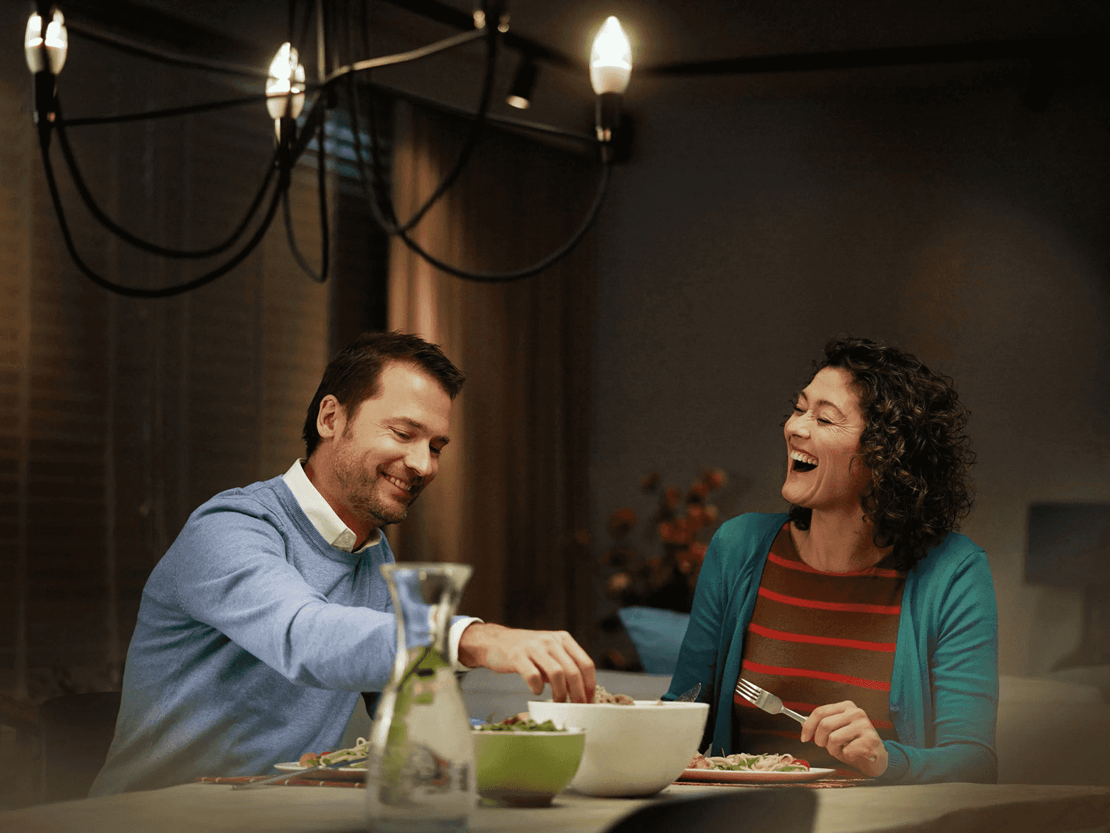 Un homme souriant et une femme riant dînent à une table avec de la nourriture et des boissons.