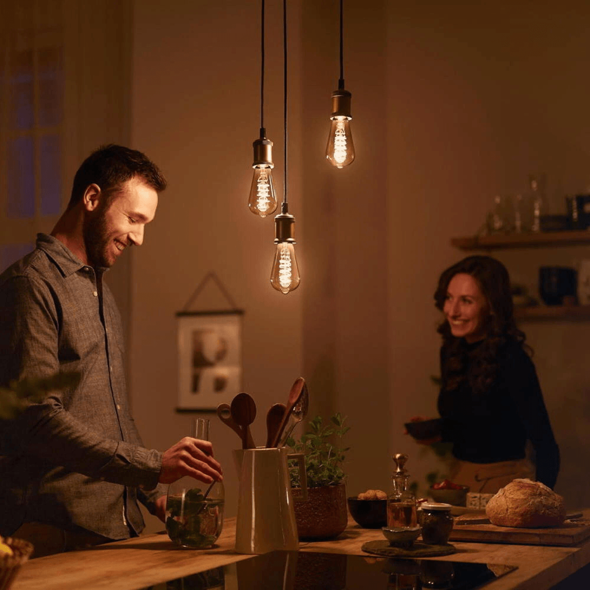 A man and woman in a kitchen under three lightbulbs. The man pours water.