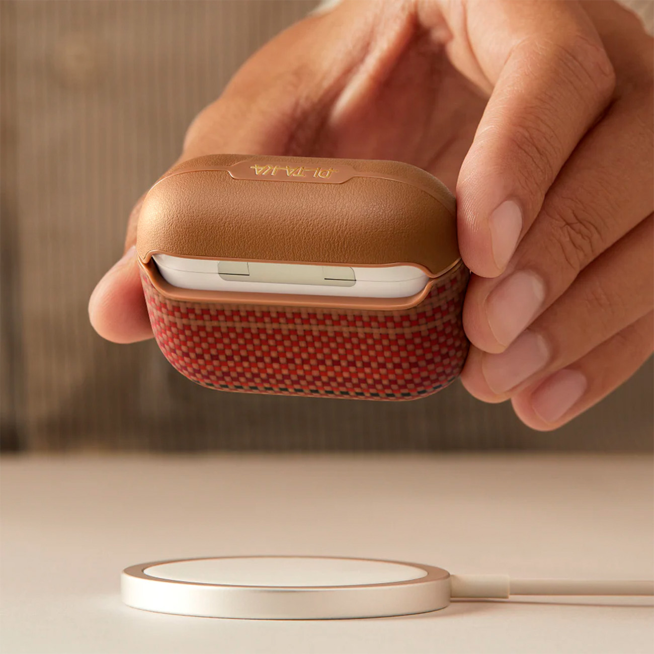 Una mano sujeta un estuche marrón y rojo para auriculares sobre una base de carga.