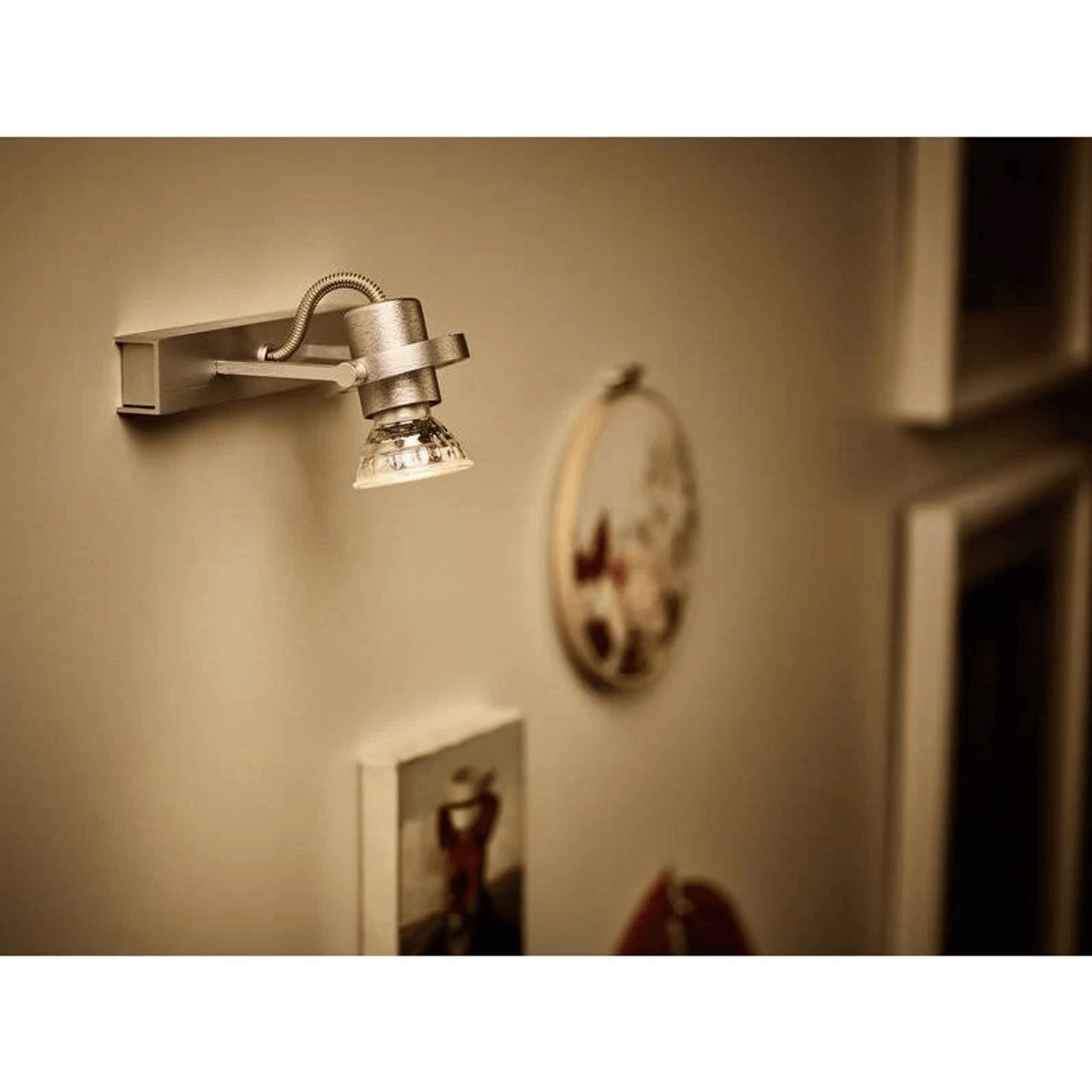 A silver wall spotlight illuminates a wall with pictures and embroidery.