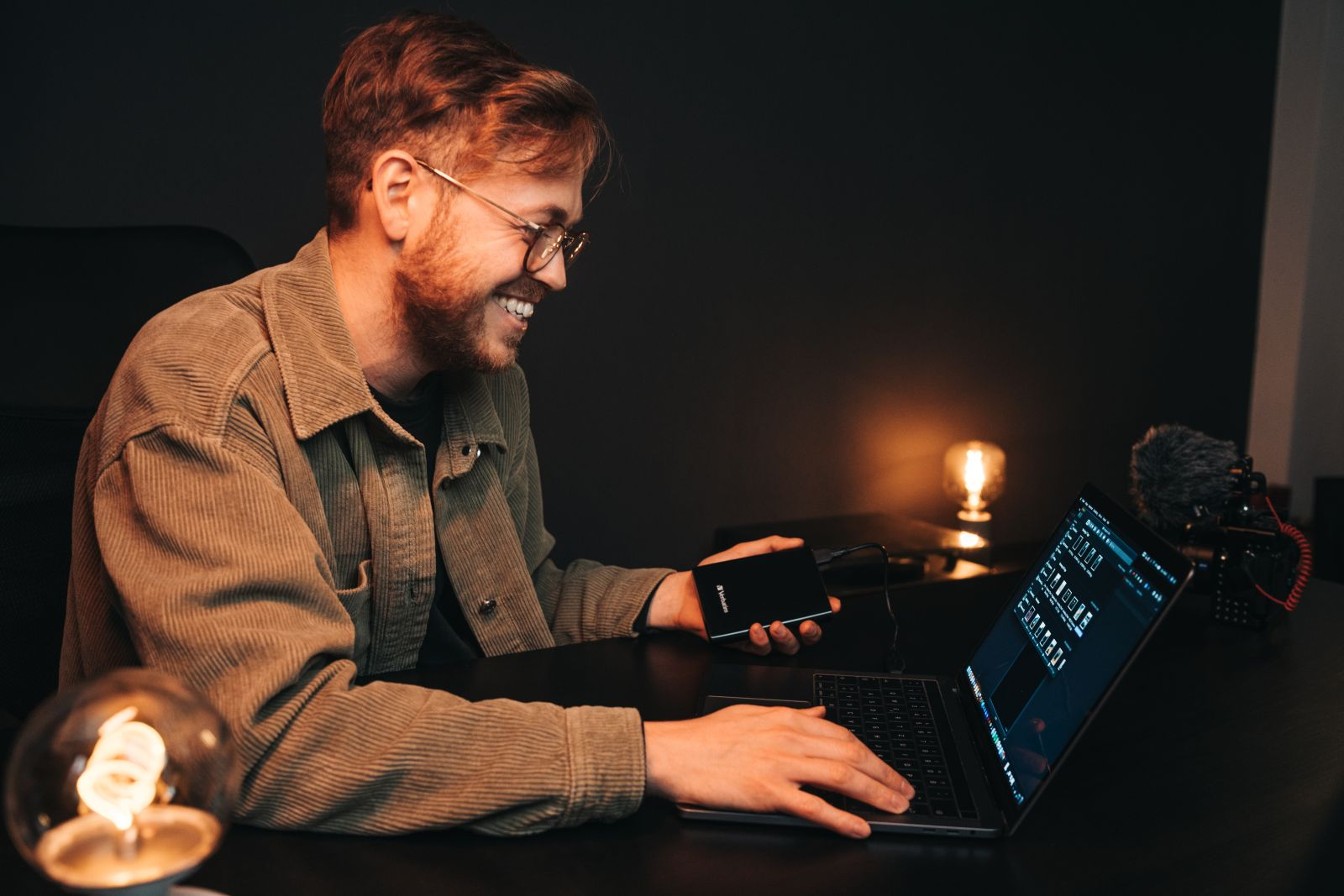 A man with glasses and a beard smiles while working on a laptop, holding an external hard drive.