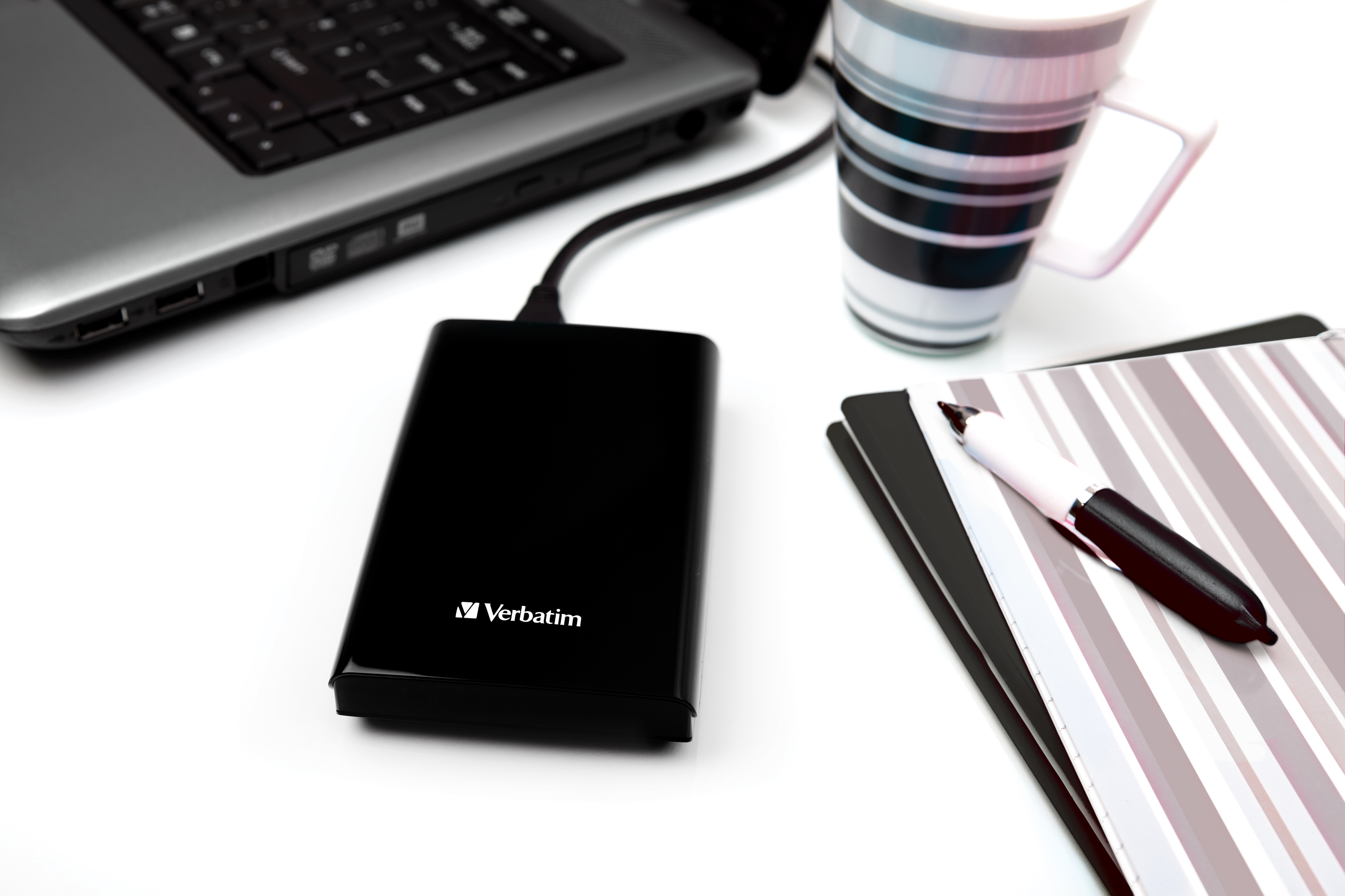 A black Verbatim external hard drive is connected to a laptop, next to a striped mug and notepad.