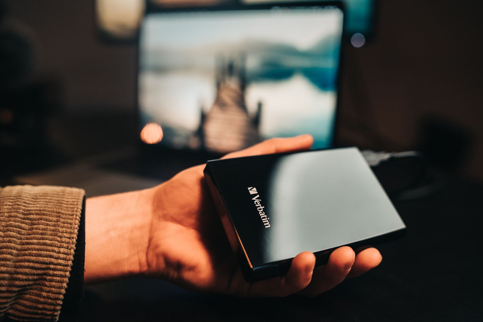 A hand holds a black Verbatim external hard drive in front of a laptop showing a landscape.
