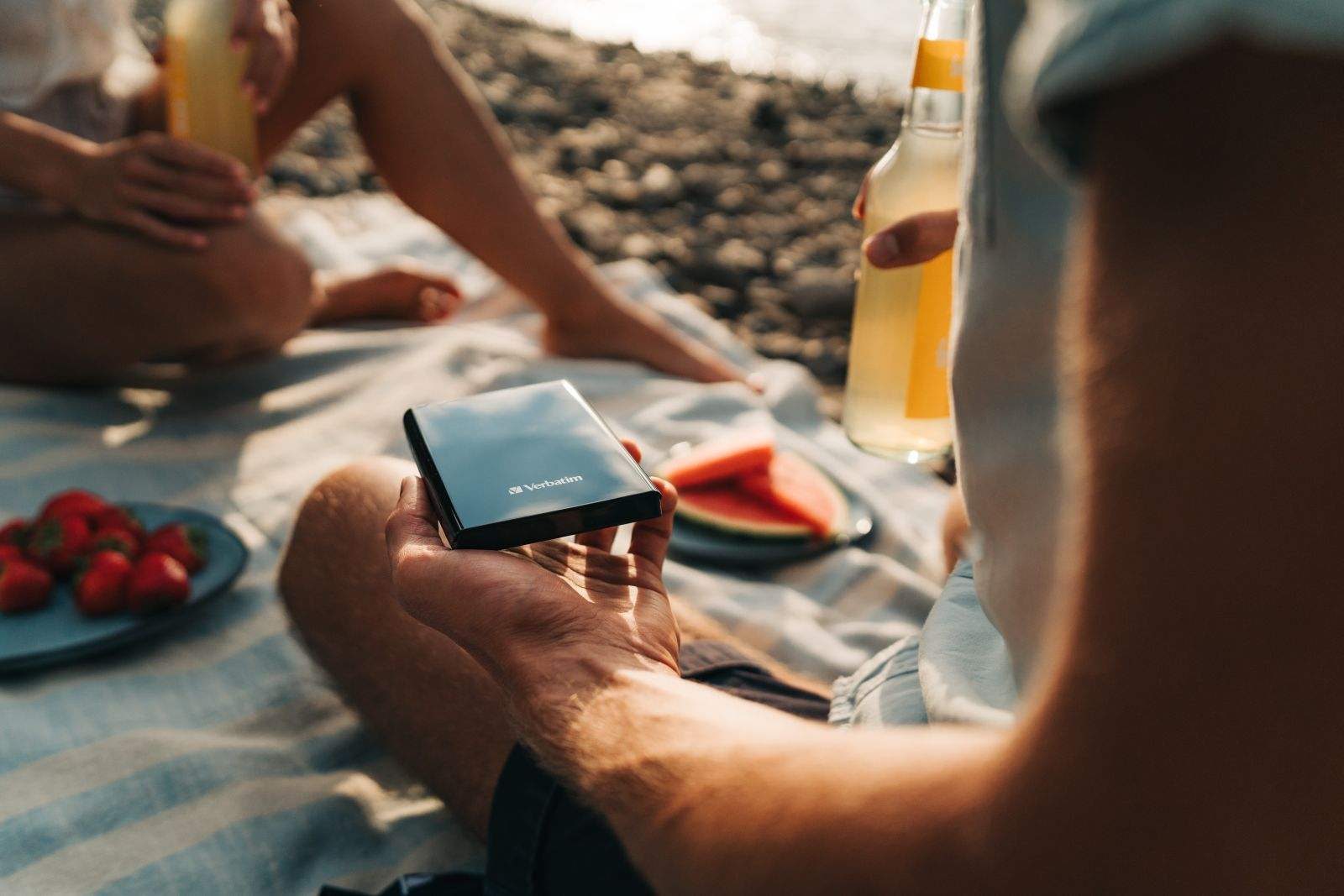 A hand holds a black hard drive on a beach blanket. Strawberries and watermelon nearby.