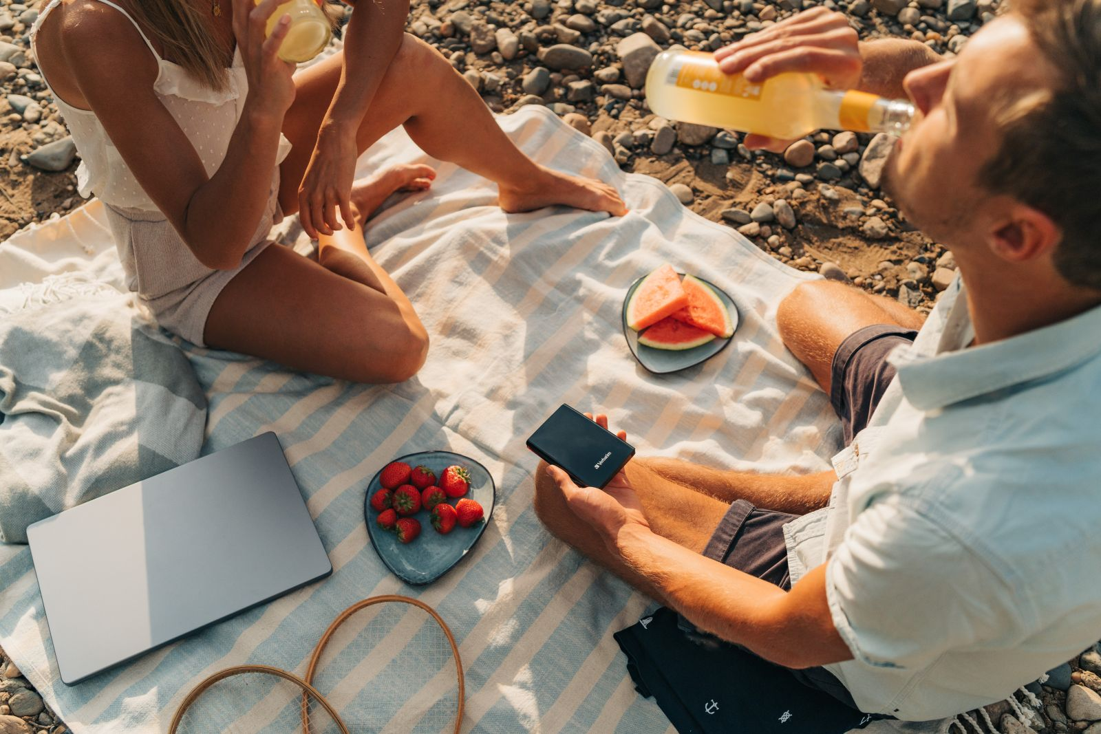People having a picnic on a beach with strawberries, watermelon, a laptop, and drinks.