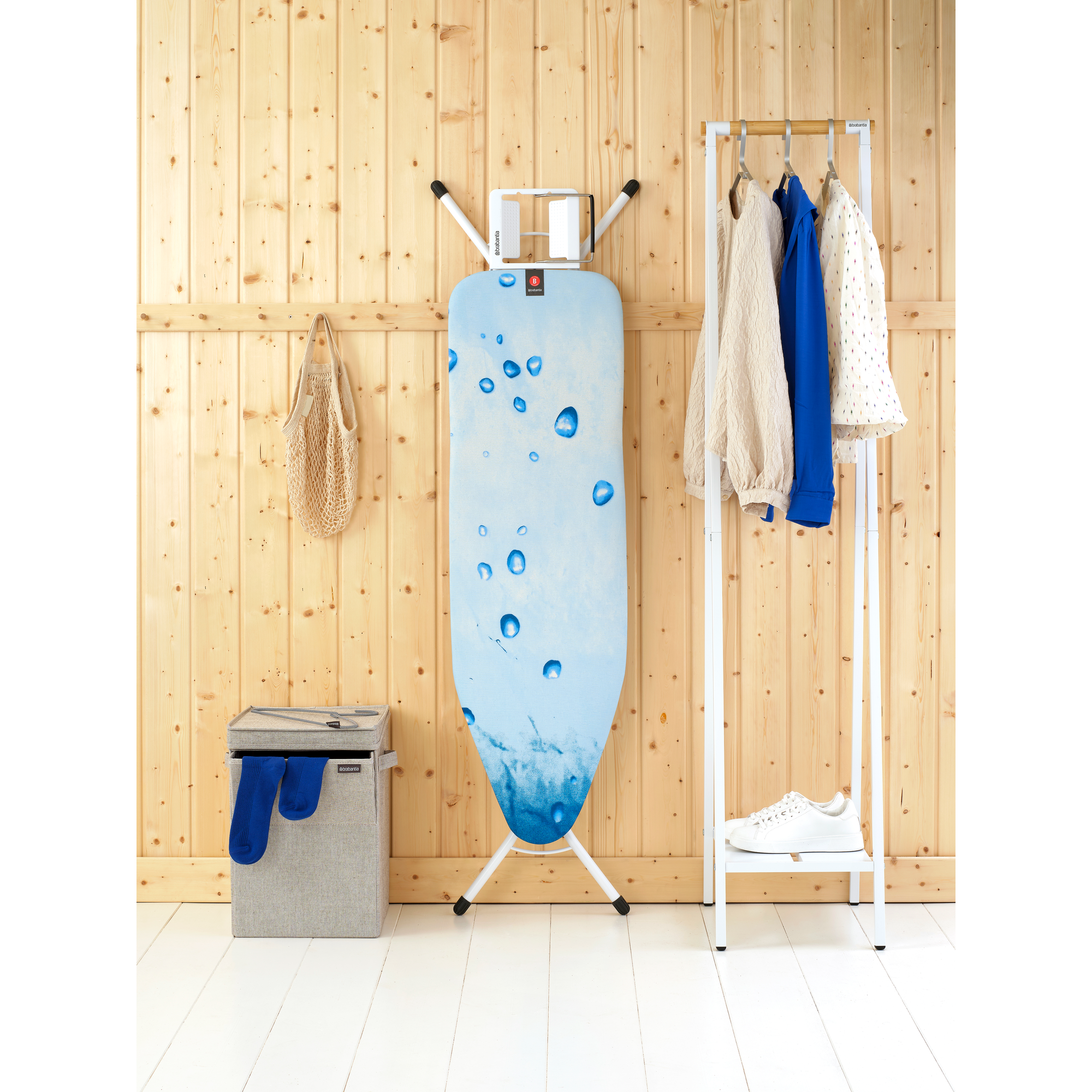 Bügeleisen, Wäschekorb, Kleiderständer mit Kleidung und Schuhen an Holzwand.