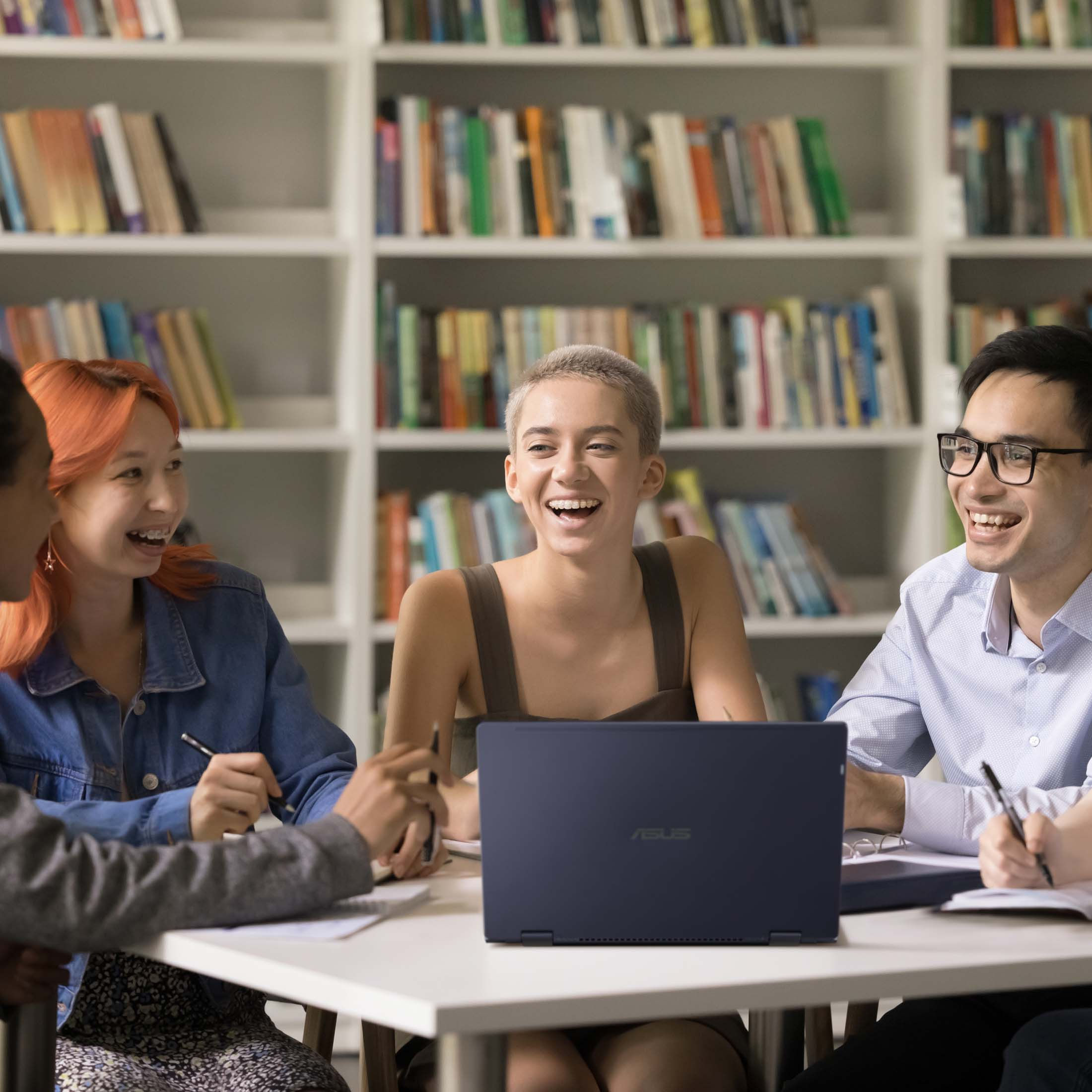 Fünf junge Leute lachen und lernen in einer Bibliothek mit Bücherregalen.