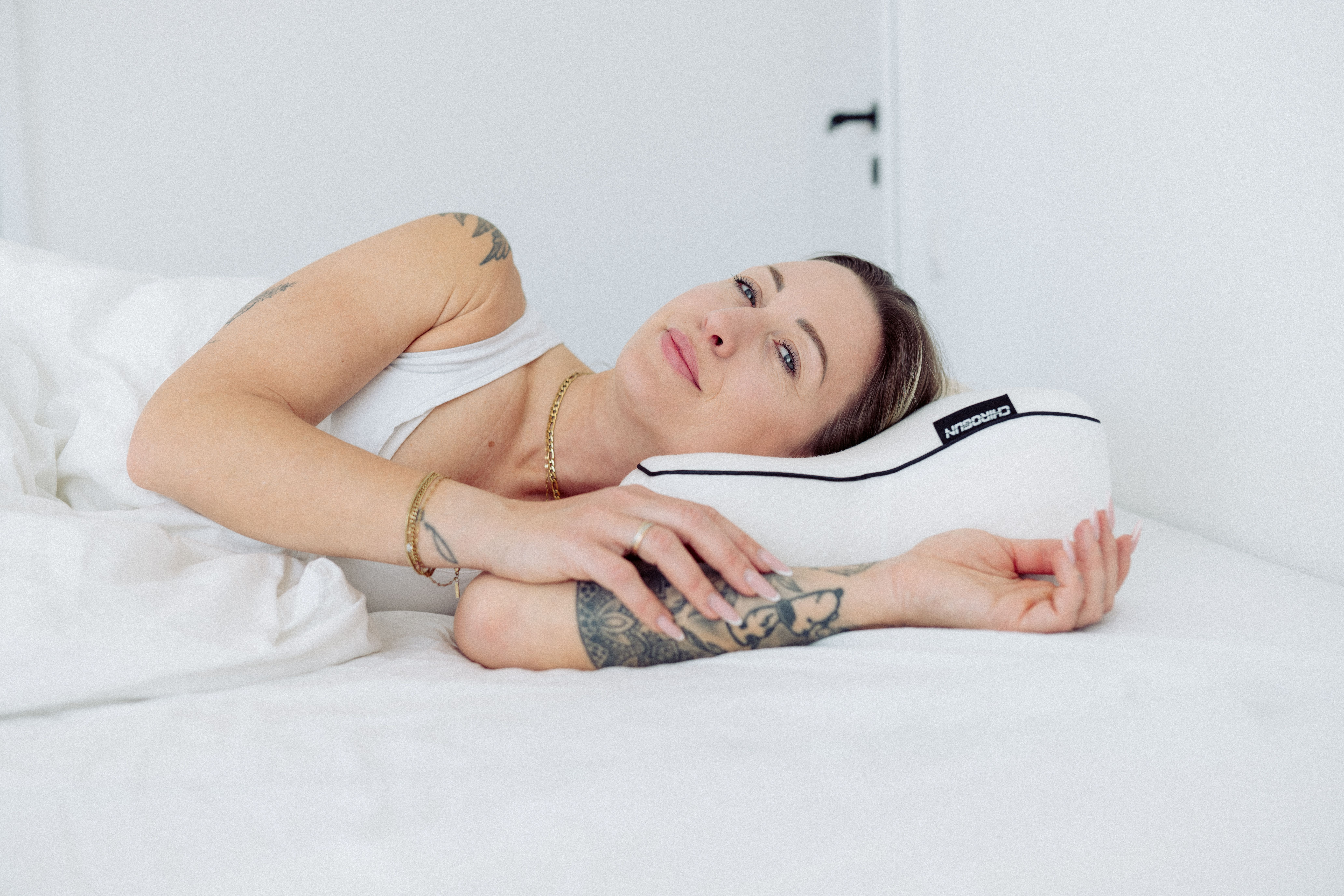 Eine Frau mit Tattoos liegt auf einem weißen Bett und hält ein Kissen.