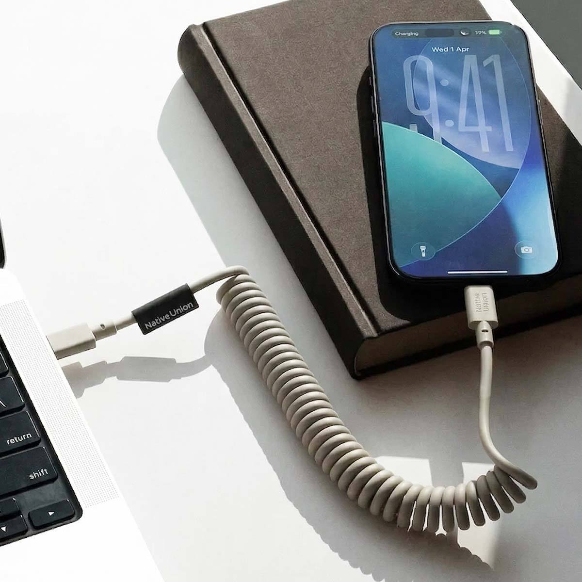 Un téléphone se charge via un câble en spirale à un ordinateur portable. Un livre est à côté.