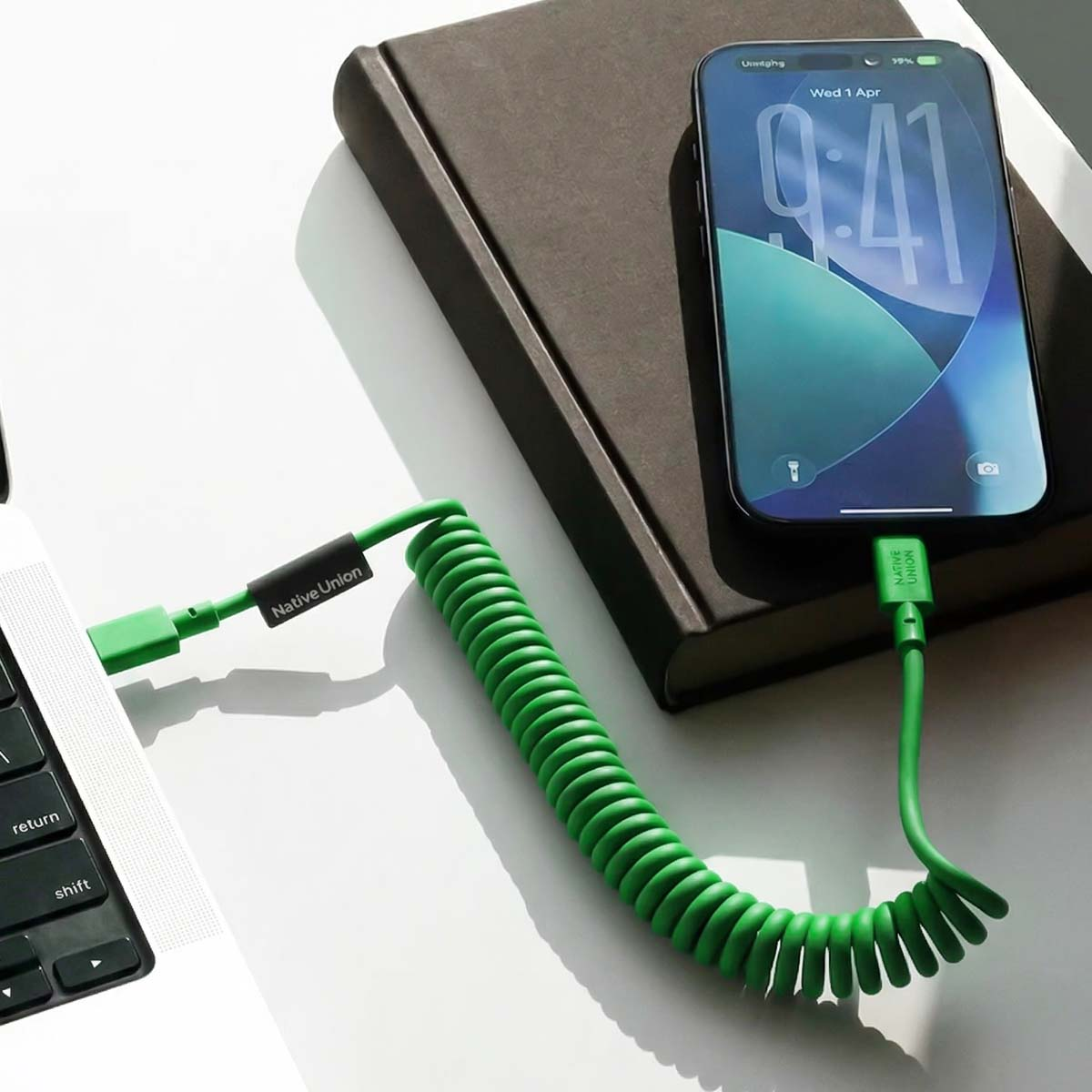 Ein grünes Spiralkabel verbindet ein Smartphone mit einem Laptop, daneben liegt ein Buch.