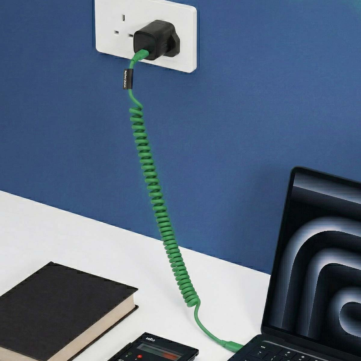 Grünes Spiralkabel verbindet schwarzen Netzstecker mit Laptop. Buch, Gerät auf Tisch.