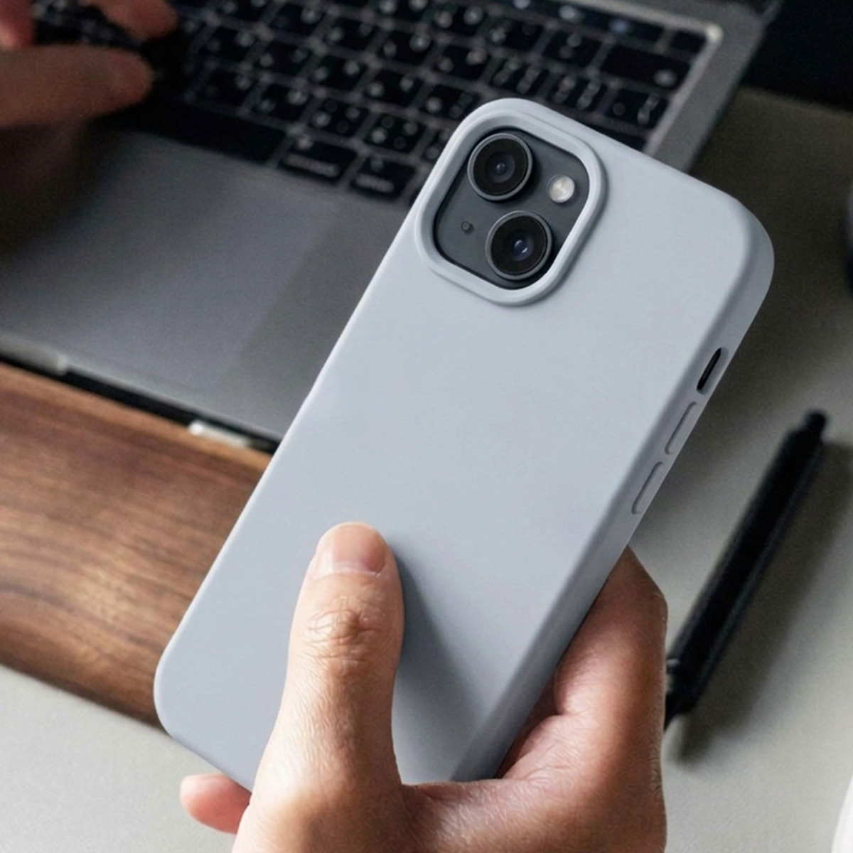Une main tient une coque grise pour iPhone avec deux caméras. Un ordinateur portable et un stylo à l'arrière.