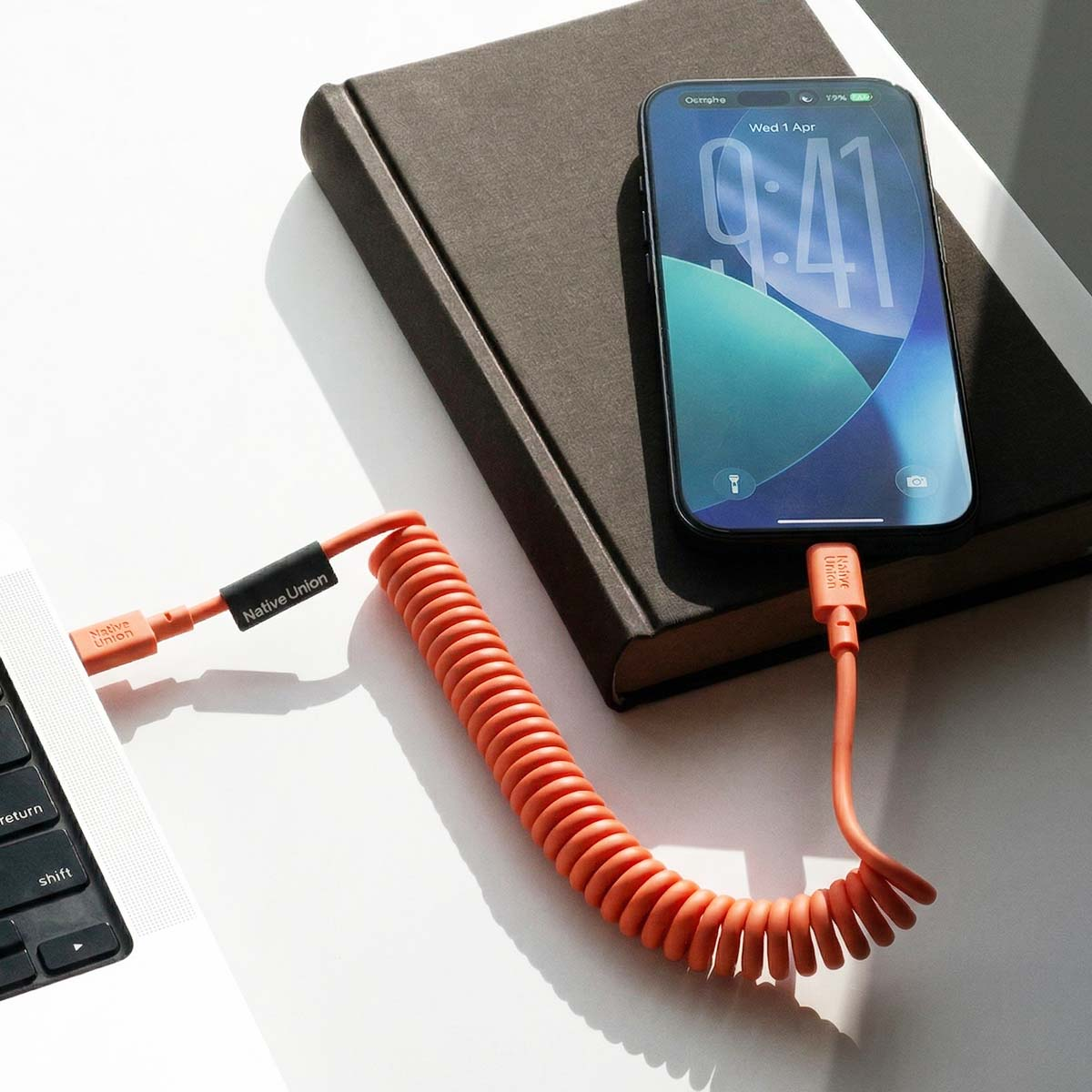 Un câble orange en spirale relie un iPhone à un ordinateur portable à côté d'un livre.