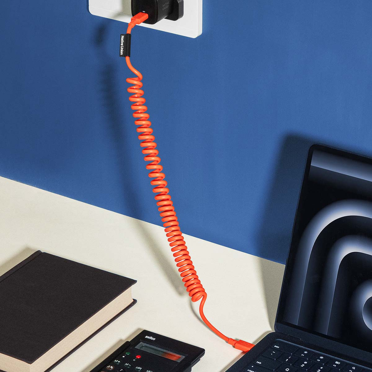 Un câble spiralé orange relie un chargeur à un ordinateur portable. Un livre et une calculatrice sont à proximité.