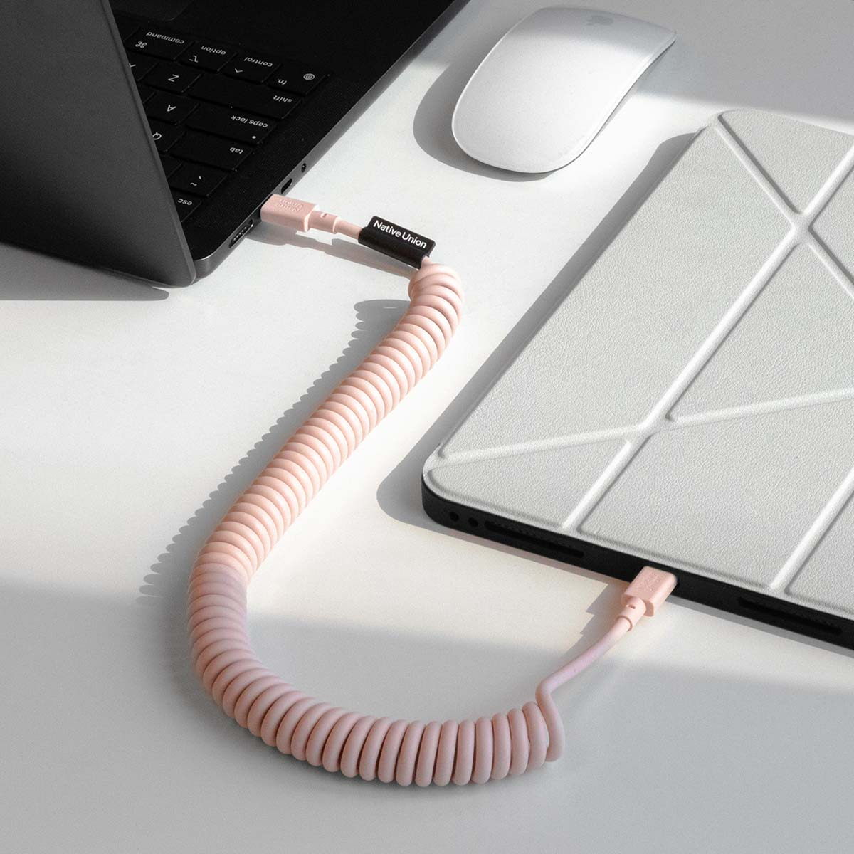 Un câble spiralé rose relie un ordinateur portable et un étui de tablette. Une souris blanche à proximité.