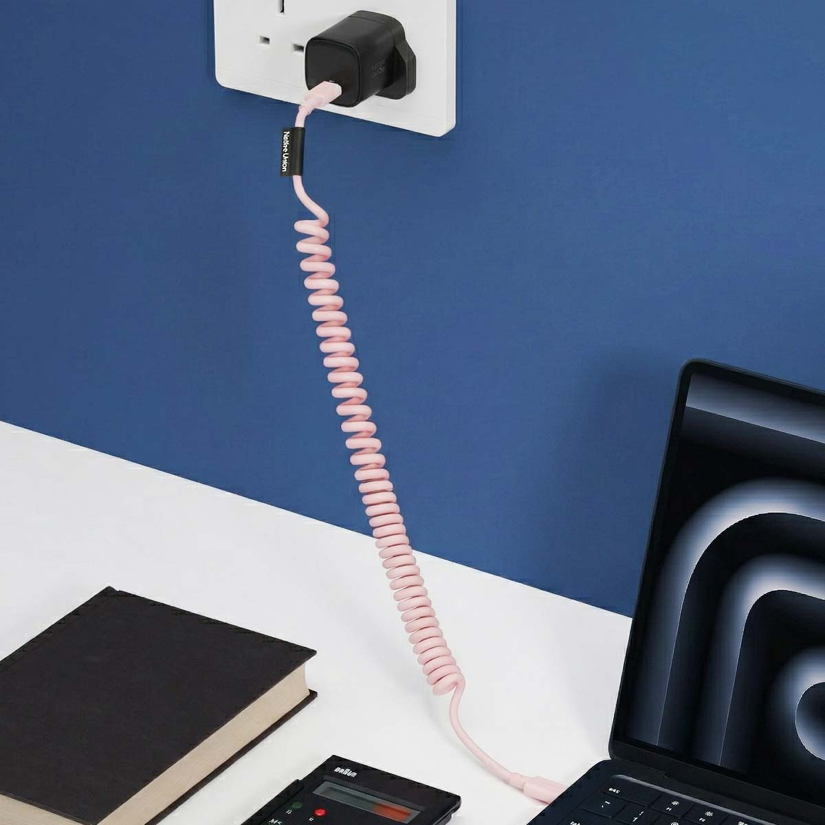 Un câble de charge rose en spirale est branché sur une prise murale et connecté à un ordinateur portable. Un livre et une calculatrice sont à proximité.