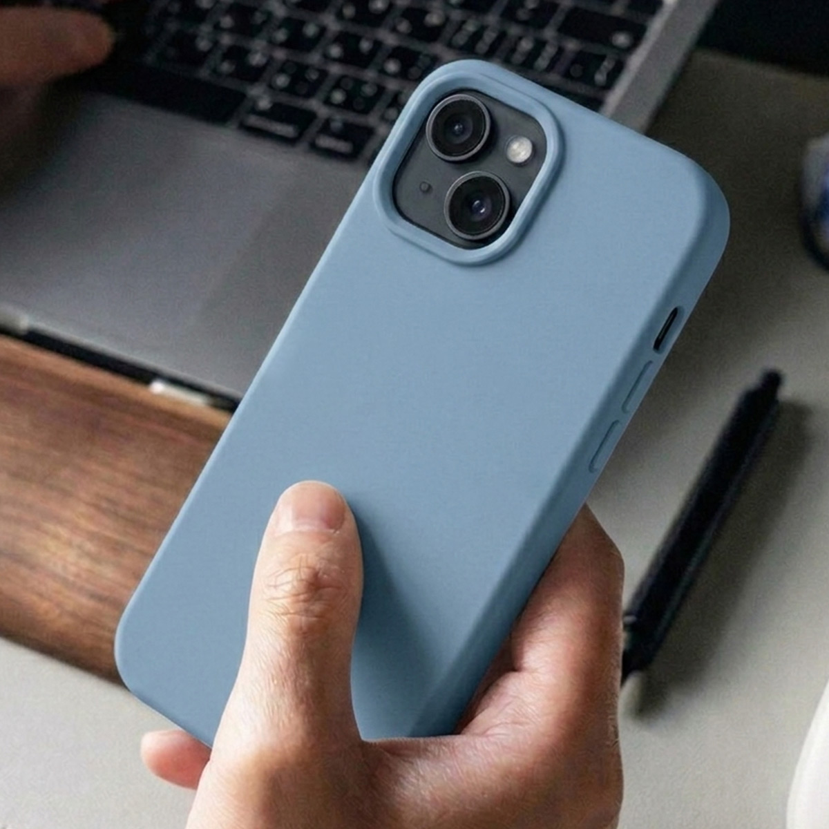 A hand holds a blue smartphone with a camera module in front of a laptop.