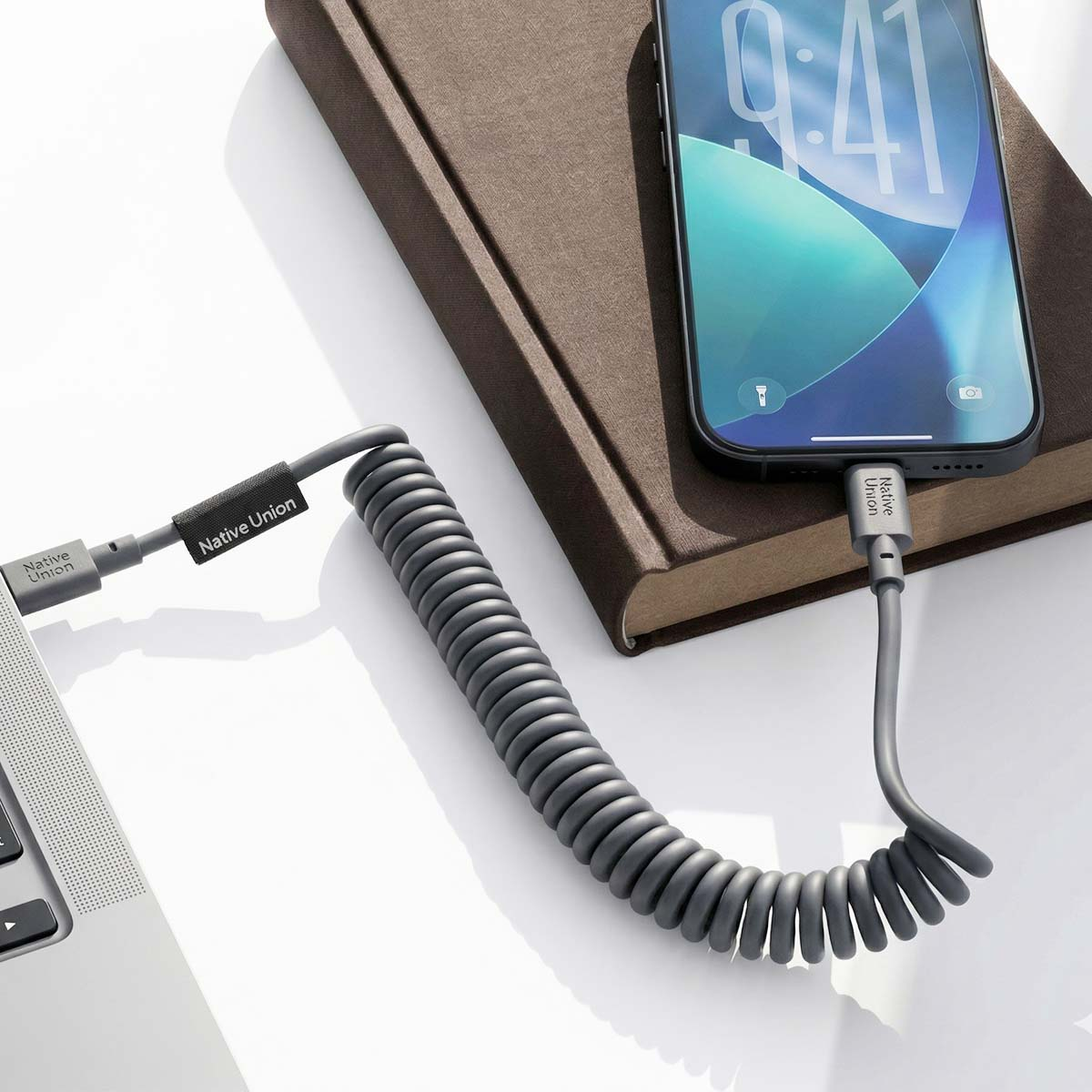 Un cavo di ricarica grigio a spirale collega un telefono a un laptop, appoggiato su un libro.