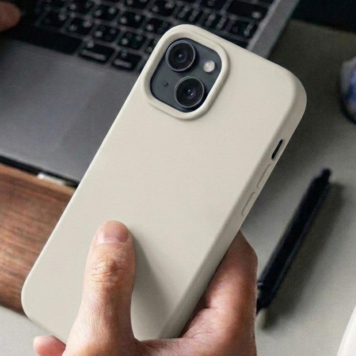 Eine Hand hält ein hellbeiges iPhone-Gehäuse vor einem Laptop und Stift.