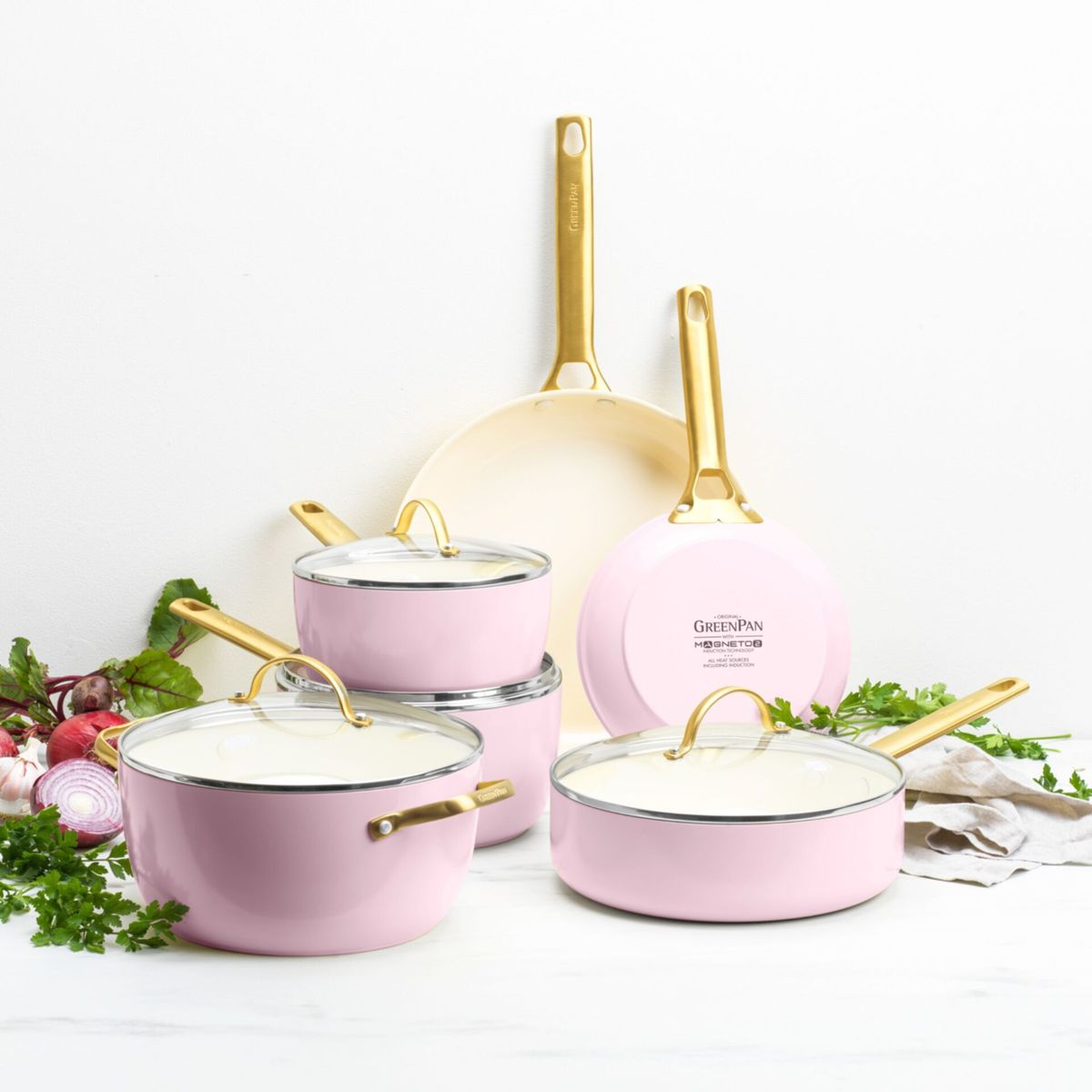 Rosa Kochgeschirr mit goldenen Griffen, daneben Gemüse und Kräuter.