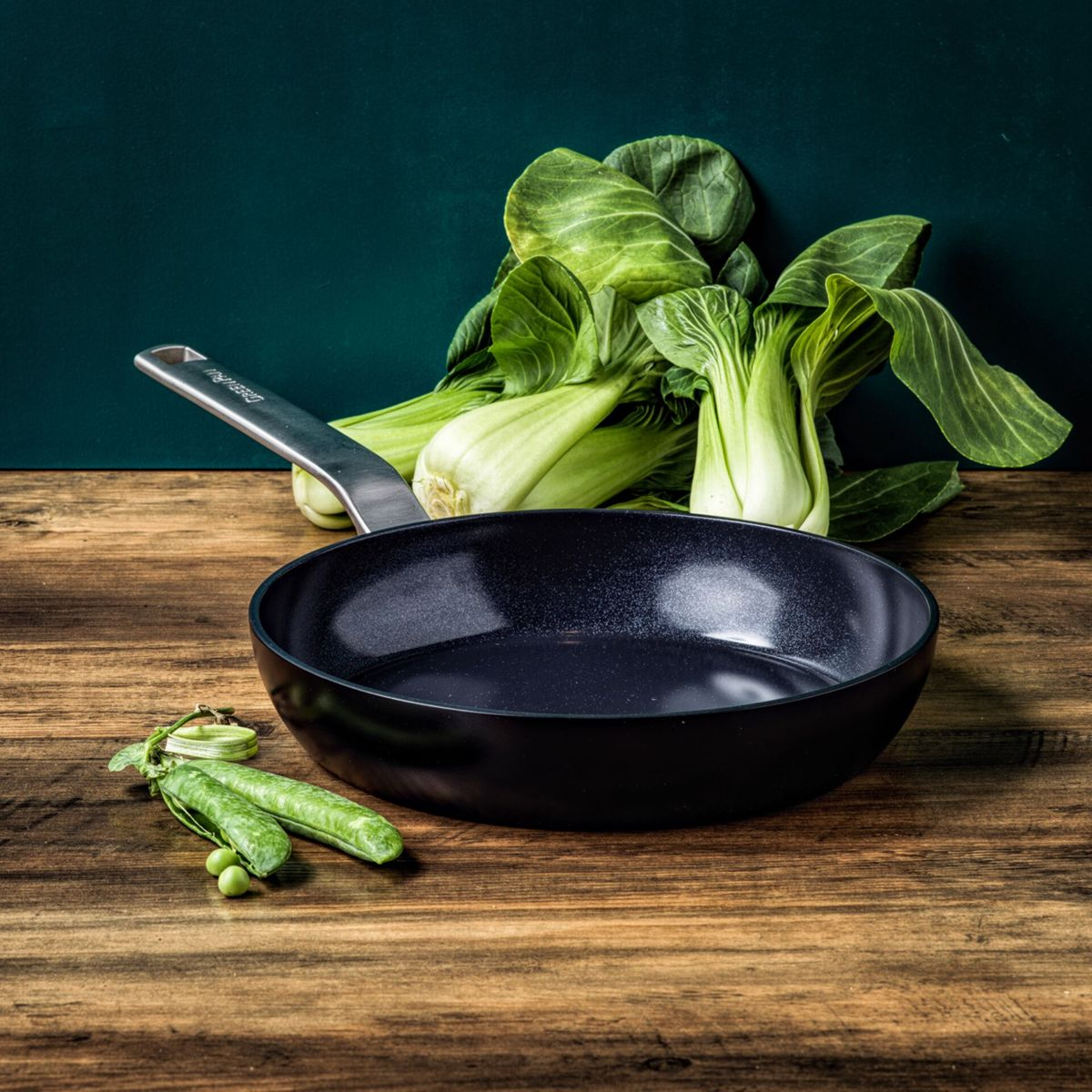 Eine schwarze Bratpfanne mit Pak Choi und Zuckerschoten auf Holztisch.