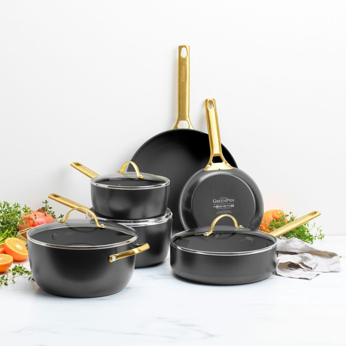 Schwarzes Kochgeschirr mit goldenen Griffen, daneben Obst und Kräuter.
