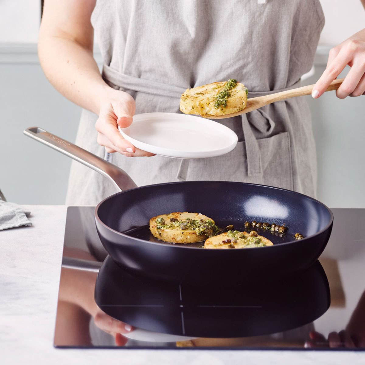 Eine Person in einer Schürze legt gebratenes Essen mit Kräutern auf einen Holzlöffel über eine Pfanne.