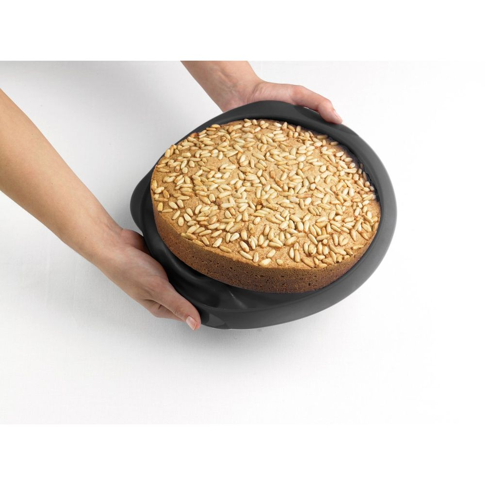 Ein runder Kuchen mit Pinienkernen in einer schwarzen Form wird von zwei Händen gehalten.