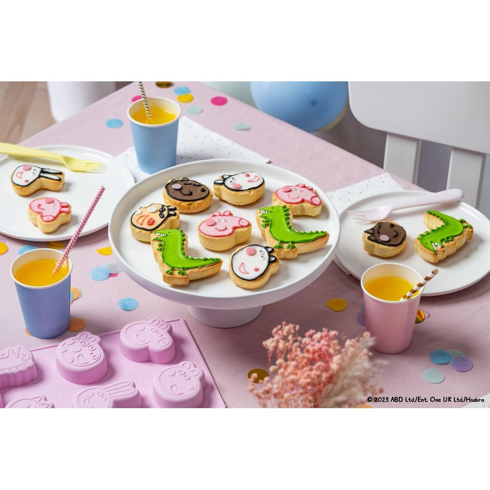 Kekse in Tierform auf Kuchenständer, Becher mit Saft und Formen auf rosafarbenem Tisch.