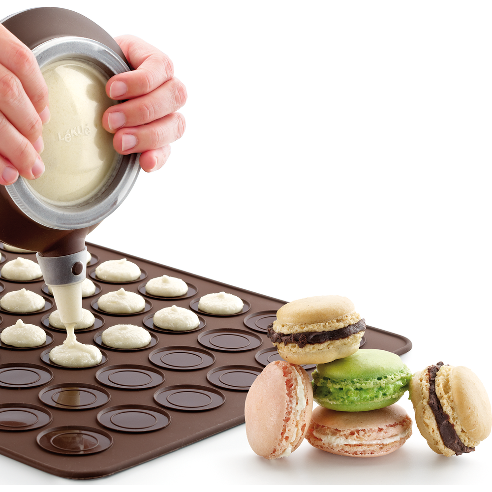 Mains remplissent moules à macarons sur tapis silicone avec pâte; macarons finis à côté.