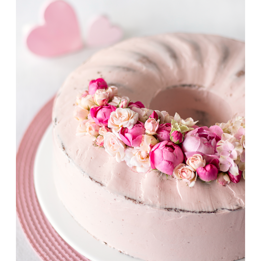 Ein rosafarbener Kranzkuchen mit rosa Buttercreme und Blumen. Zwei Herzen im Hintergrund.