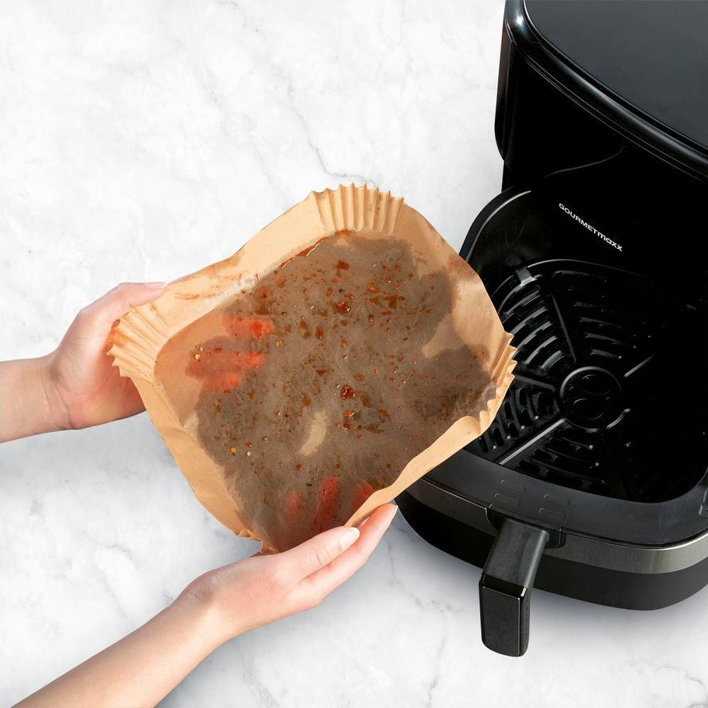 Hände halten braunes Papier mit Essensresten neben einer schwarzen Heißluftfritteuse.