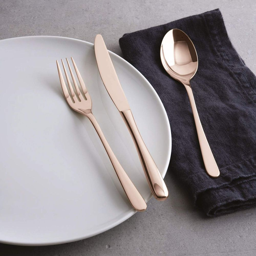 Couverts en or rose sur une assiette blanche et un tissu sombre.