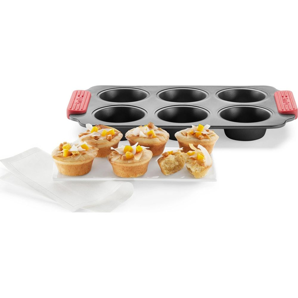 Sechs kleine Cupcakes mit Kokosnuss und Obst bedeckt stehen auf einem weißen Teller neben einer Muffinform.