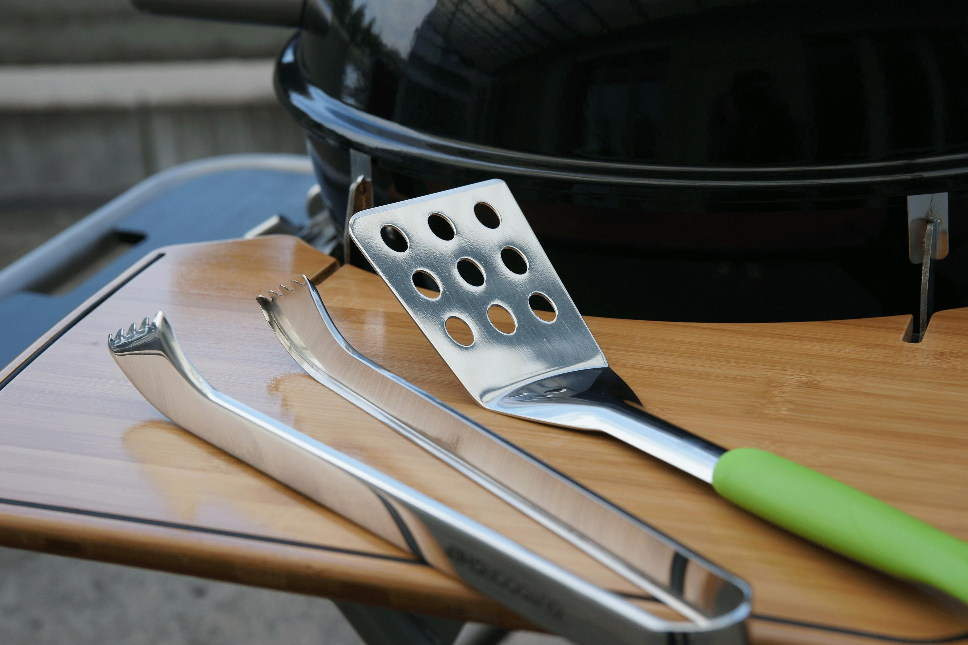 Attrezzi da barbecue su tavolo di legno davanti a un grill nero.