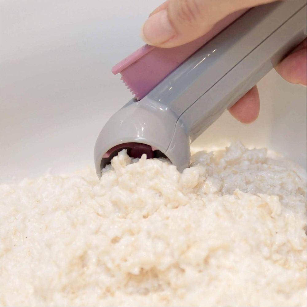 Une main utilise un appareil gris et rose pour faire des boules de riz dans du riz blanc.