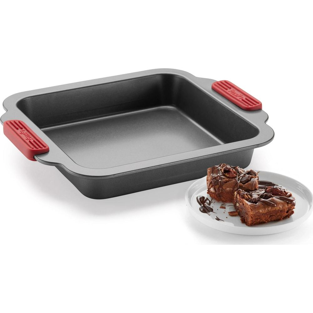 Un moule à gâteau carré avec des poignées rouges à côté d'une assiette avec deux brownies au chocolat.