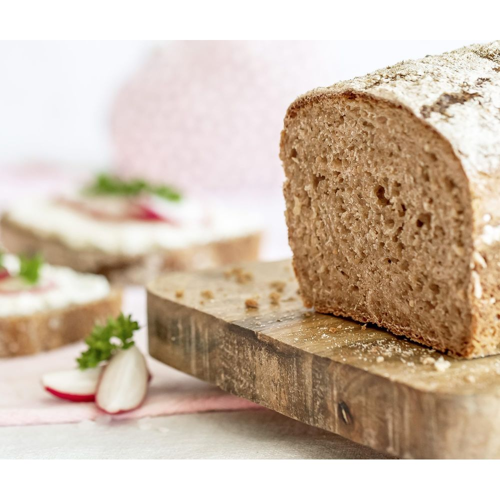 Ein Laib dunkles Brot auf einem Holzbrett, daneben Brotscheiben mit Frischkäse und Radieschen.