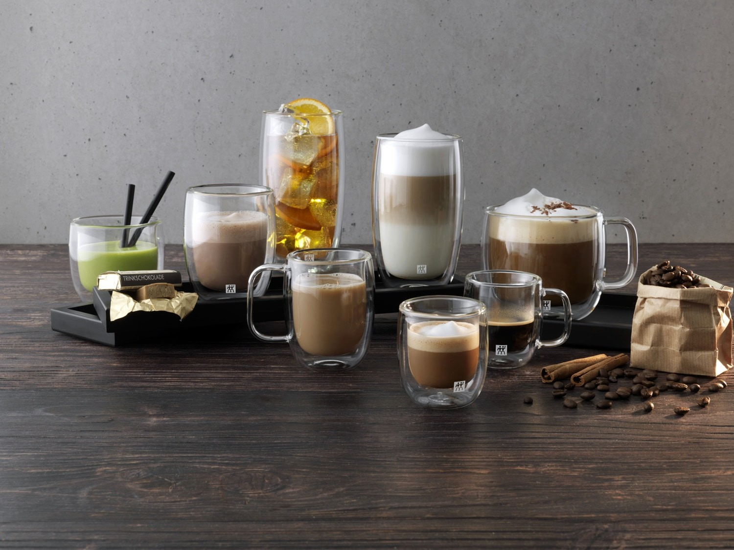 Mehrere Tassen Kaffeegetränke, ein grüner Drink, ein Orangengetränk und Schokolade auf einem Holztisch.