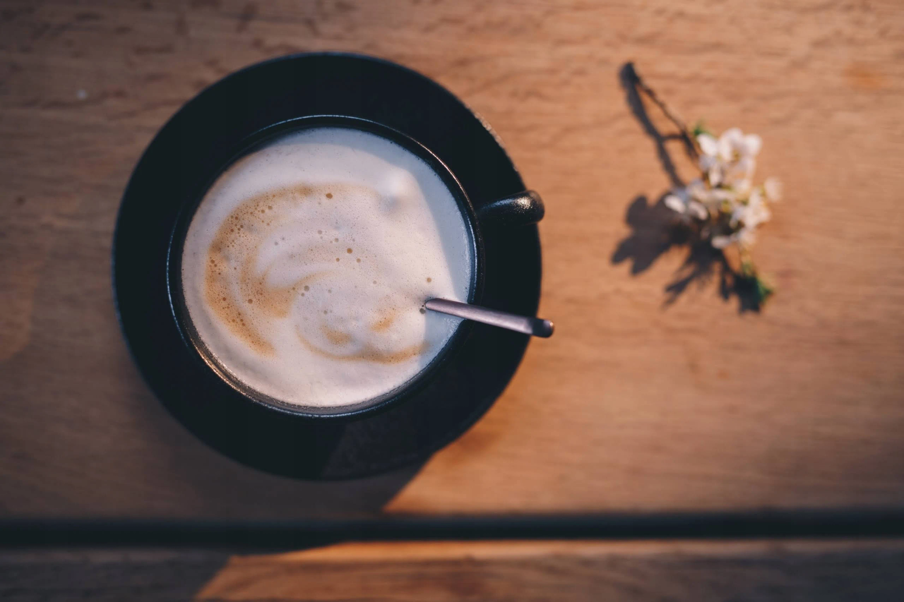Una tazza nera di caffè schiumoso con un cucchiaino su un tavolo di legno, accanto a piccoli fiori bianchi.