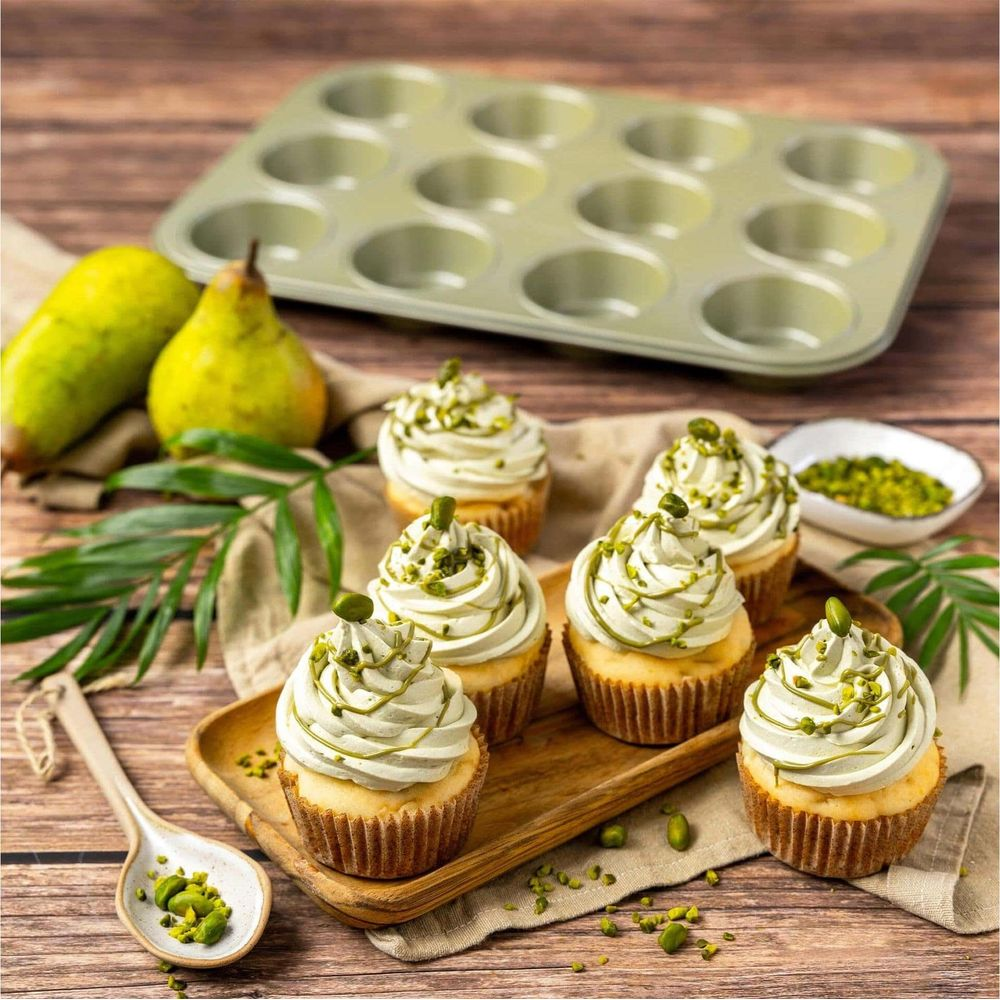 Sechs Cupcakes mit hellgrüner Glasur und Pistazien auf einem Holzteller, daneben Birnen und Backform.