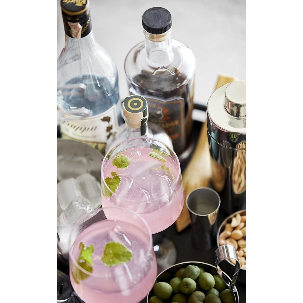 Due cocktail rosa con ghiaccio e menta, bottiglie e attrezzi da bar su un vassoio.