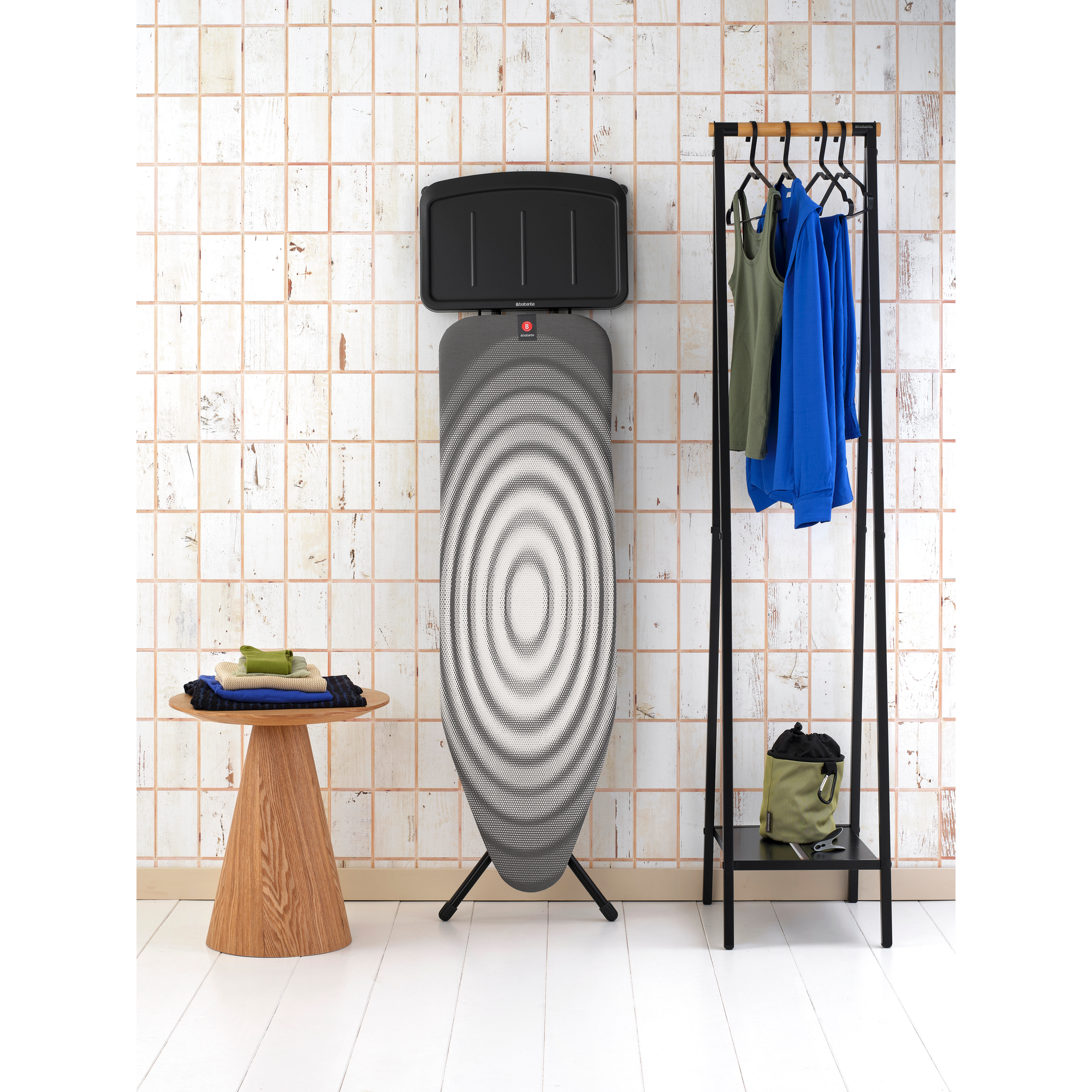 Ein Bügelbrett steht neben einem Kleiderständer mit Kleidung und einem Holztisch.