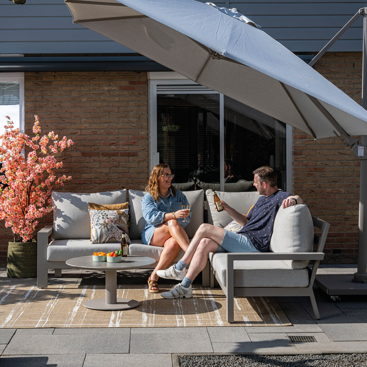 A man and woman sit on an outdoor sofa under an umbrella, drinking beer.