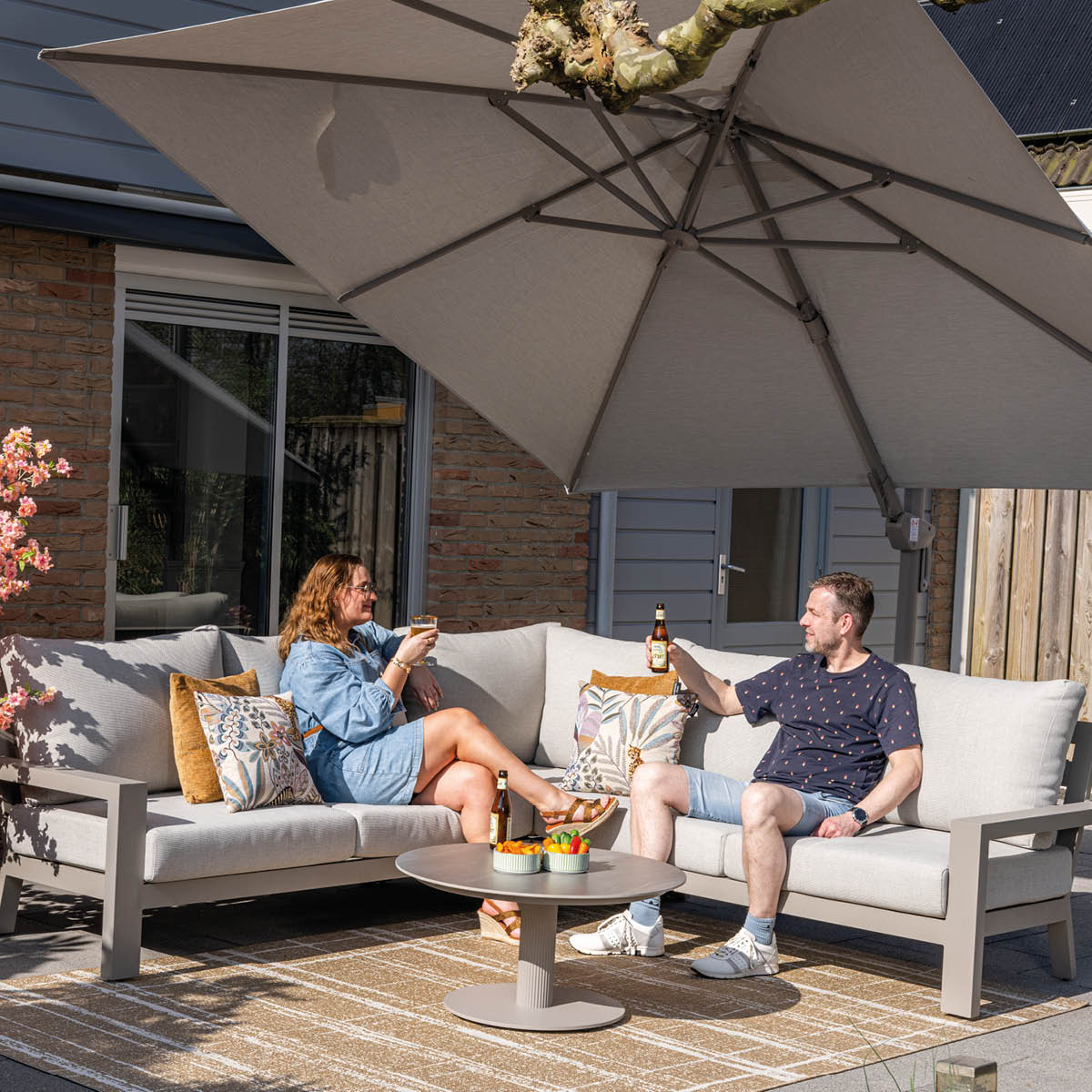 A couple enjoys drinks under a large umbrella on an outdoor sectional sofa.