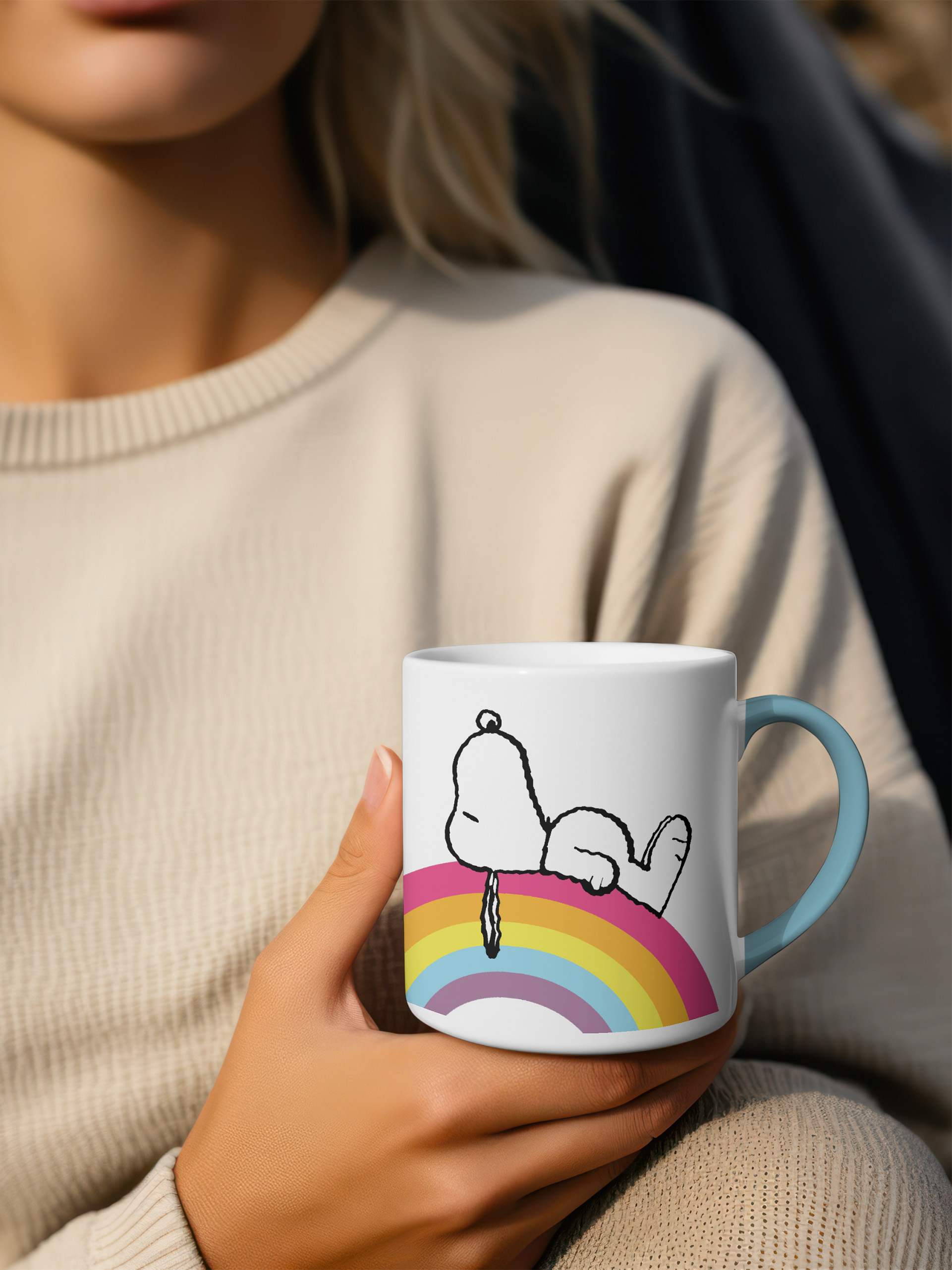 Eine weiße Tasse mit Snoopy auf einem bunten Regenbogen und einem blauen Henkel.