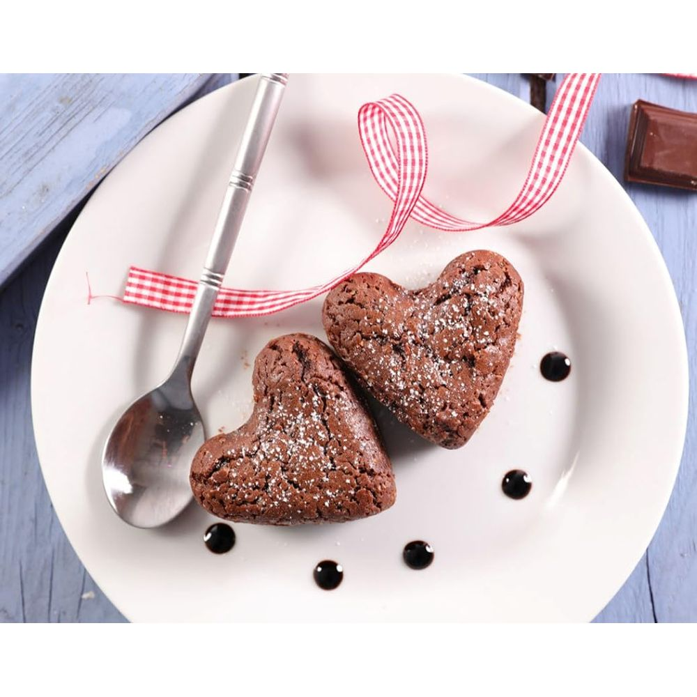 Dwa brownie w kształcie serca posypane cukrem pudrem i sosem czekoladowym na talerzu.