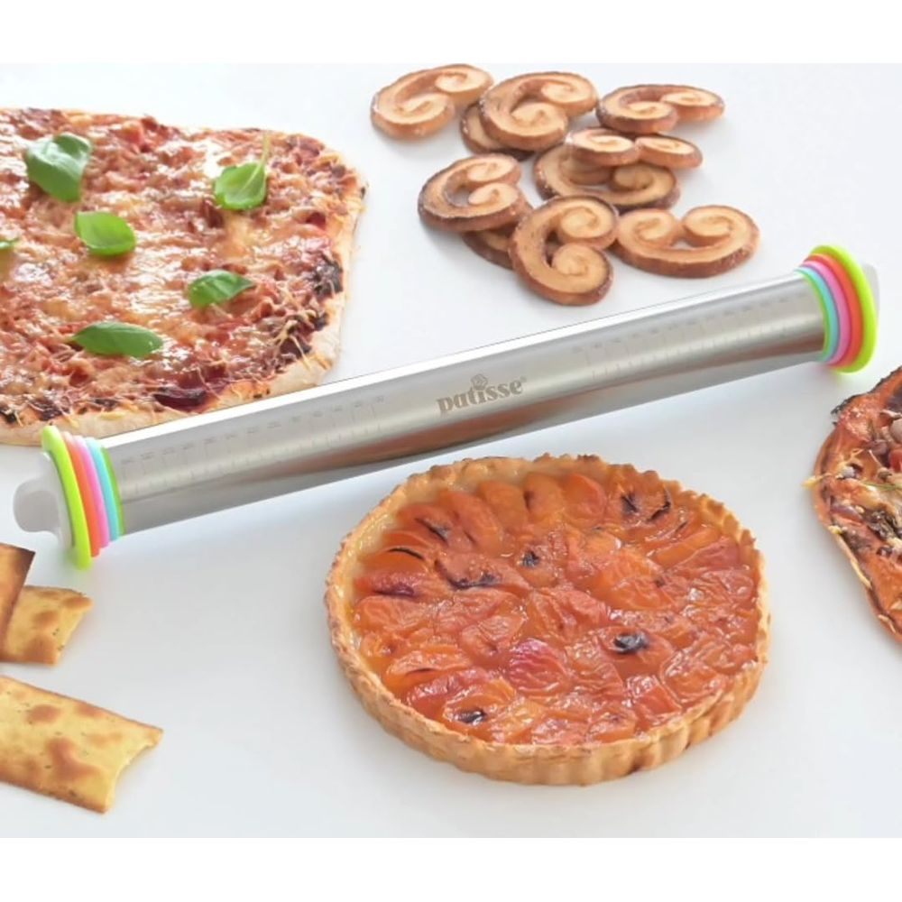 Wałek ze stali nierdzewnej z kolorowymi pierścieniami leży na białej powierzchni z pizzą i tartami.