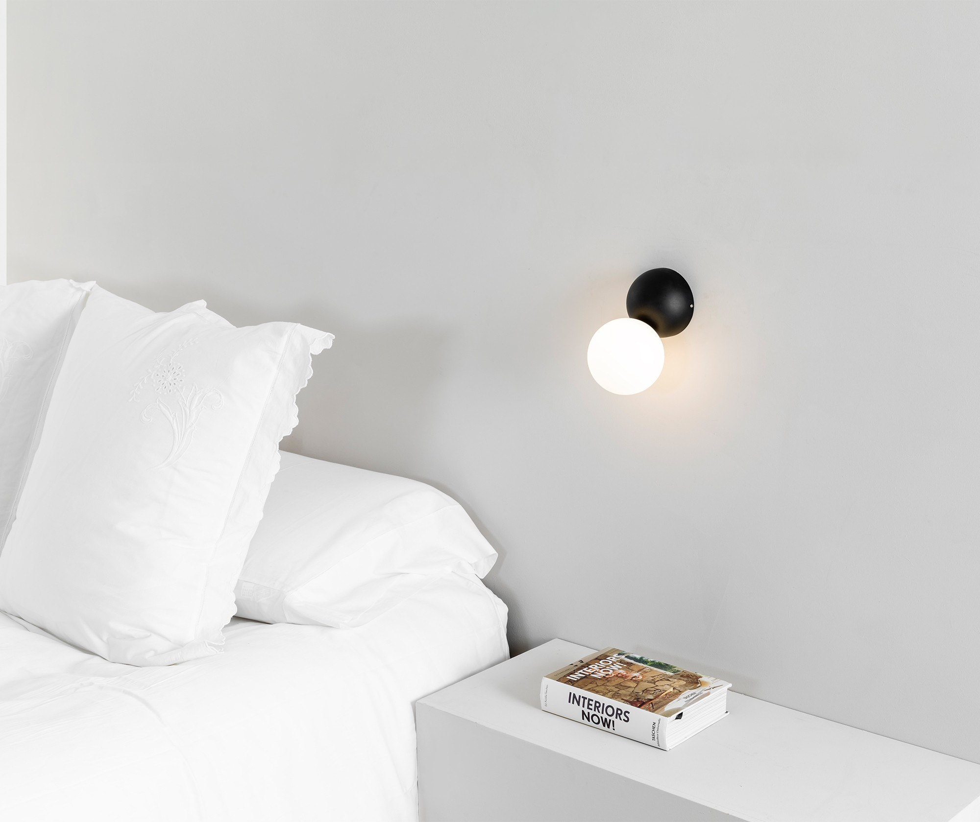 Un comodino con un libro accanto a un letto con cuscini bianchi e una lampada da parete.